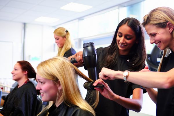 Teacher Helping Student training to become a hair dresser. The student is blow drying a woman's hair whilst the teacher watches and shows her the technique for the angle.